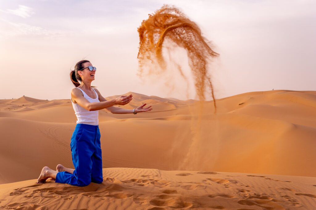 A woman in her 30s playing with sand in a desert, wearing blue pants and a white tank top, enjoying a sunny day.